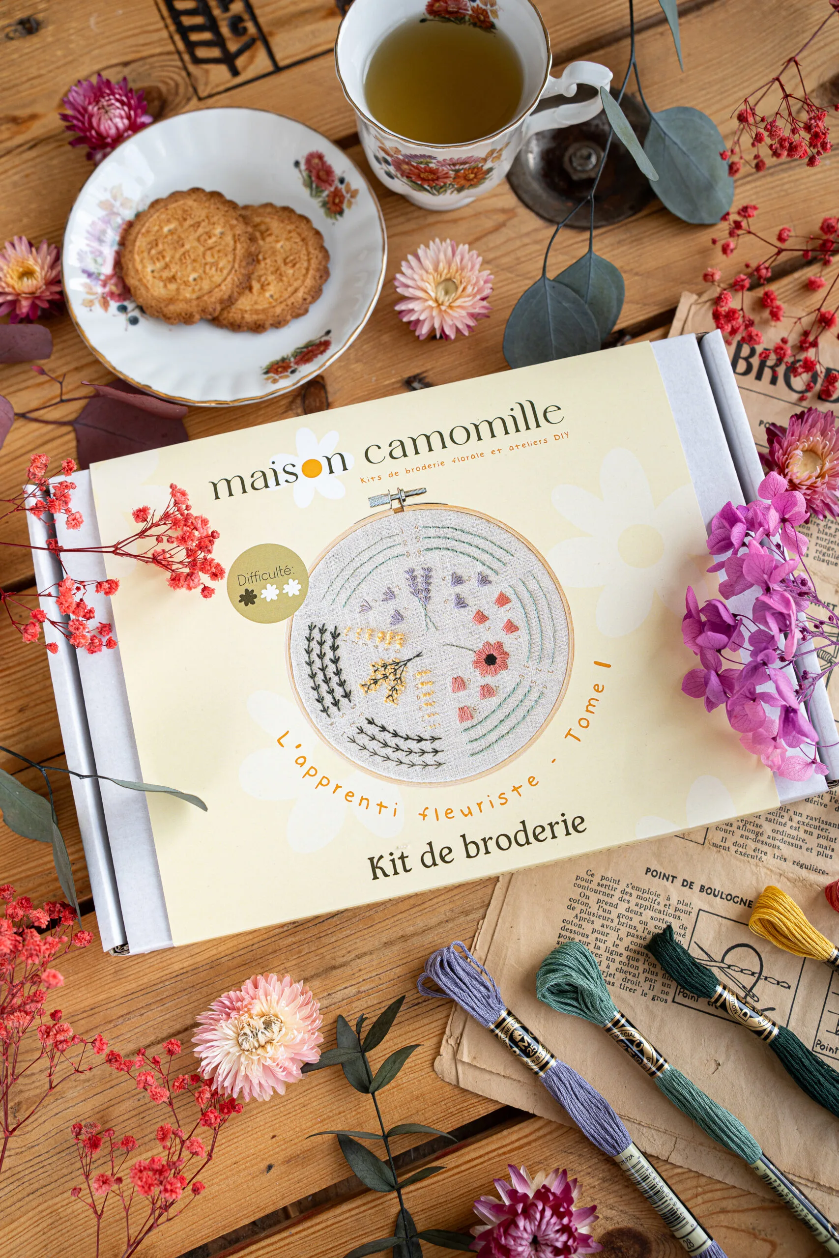 Kit broderie "L'apprenti fleuriste" Tome 1 dans son emballage, posé sur une table près d'une tasse de thé et d'une coupelle de petits gateaux, prêt à être brodé